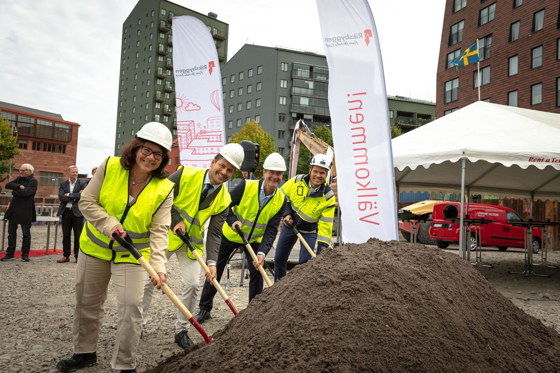Personer i gula reflexvästar och vita hjälmar använder spadar för att gräva framför en jordhög på en byggarbetsplats, omgivna av byggnader och skyltar med texten "Välkommen!