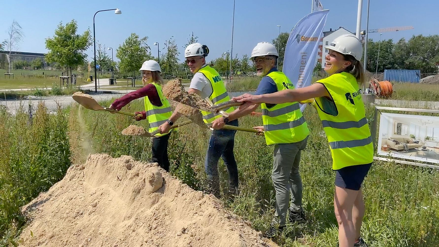 Flera personer iklädda reflexvästar och byggarbetsplats-hjälmar använder spadar för att gräva i en hög med sand på en gräsyta, med byggutrustning och flaggor i bakgrunden