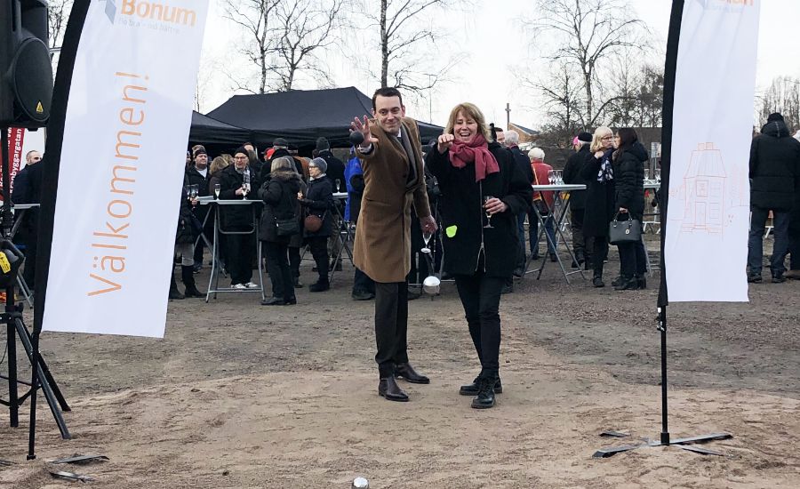 Två personer står framför en utomhuspublik mellan två vepor med texten "Välkommen!" på en marknads- eller evenemangsplats