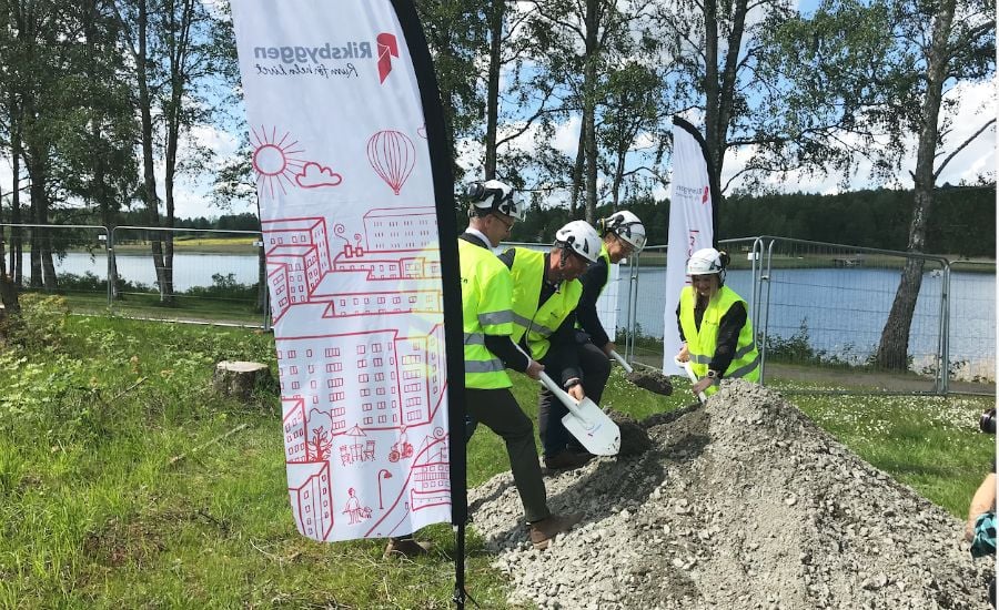Tre personer i reflexvästar och hjälmar gräver med spadar vid en grushög nära en sjö, med rollups med illustrationer i bakgrunden