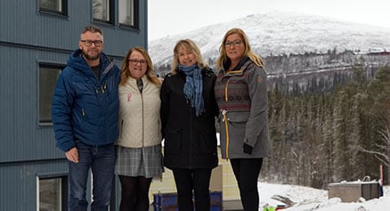 Fyra personer står framför en byggnad, med snötäckta berg och skog i bakgrunden