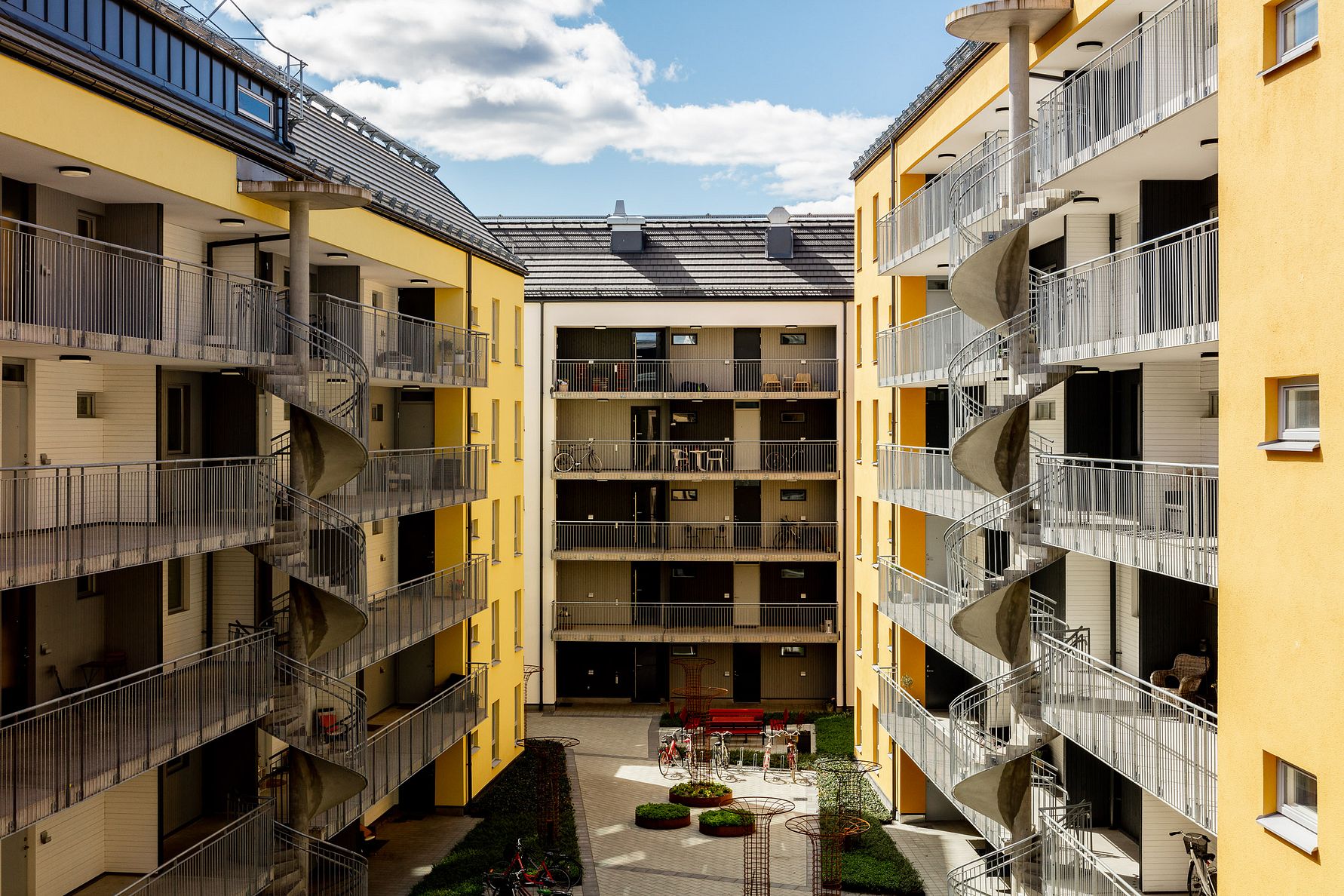 Innergård med moderna lägenhetsbyggnader, gula fasader, balkonger och spiraltrappor, omgiven av blå himmel och moln