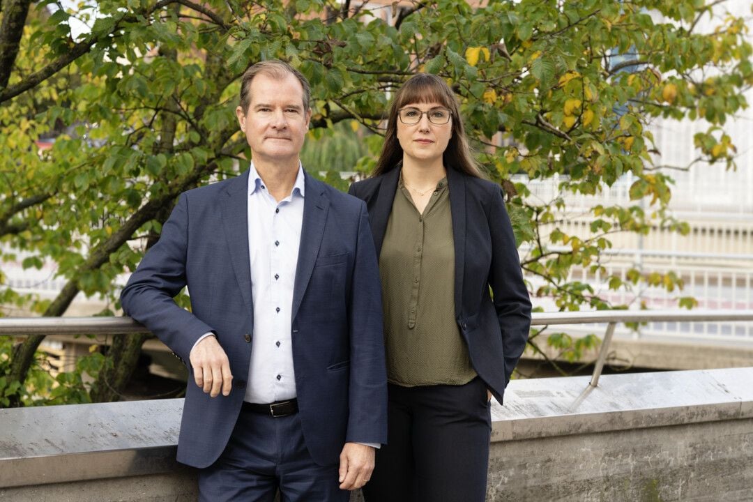 Två personer i formella kläder står utomhus framför ett träd med gröna och gula blad, nära ett räcke