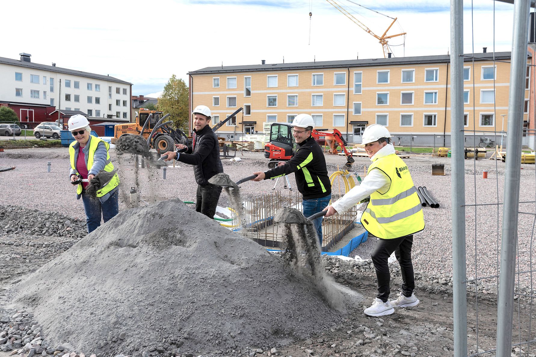 Personer i arbetskläder och hjälmar sprider grus med hjälp av ett stängsel på en byggarbetsplats, omgivna av maskiner och flerfamiljshus i bakgrunden
