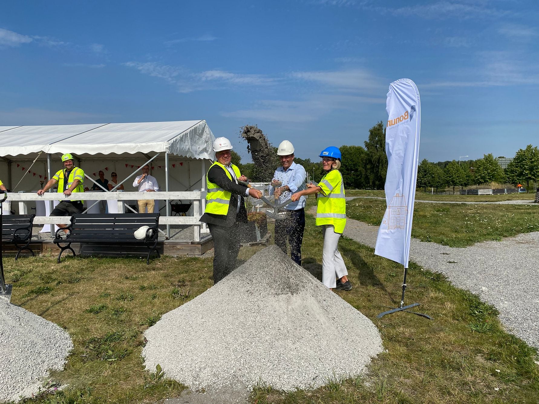 Tre personer i säkerhetskläder och hjälmar skottar grus vid en ceremoni utomhus nära ett tält och en flagga med logotyp