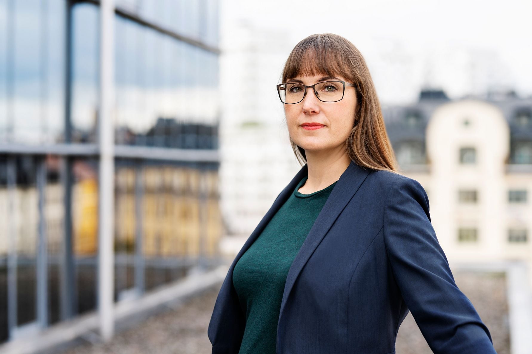 En kvinna i mörkblå kavaj och grön tröja står utomhus framför en stadsbakgrund med moderna byggnader och spegelfasader