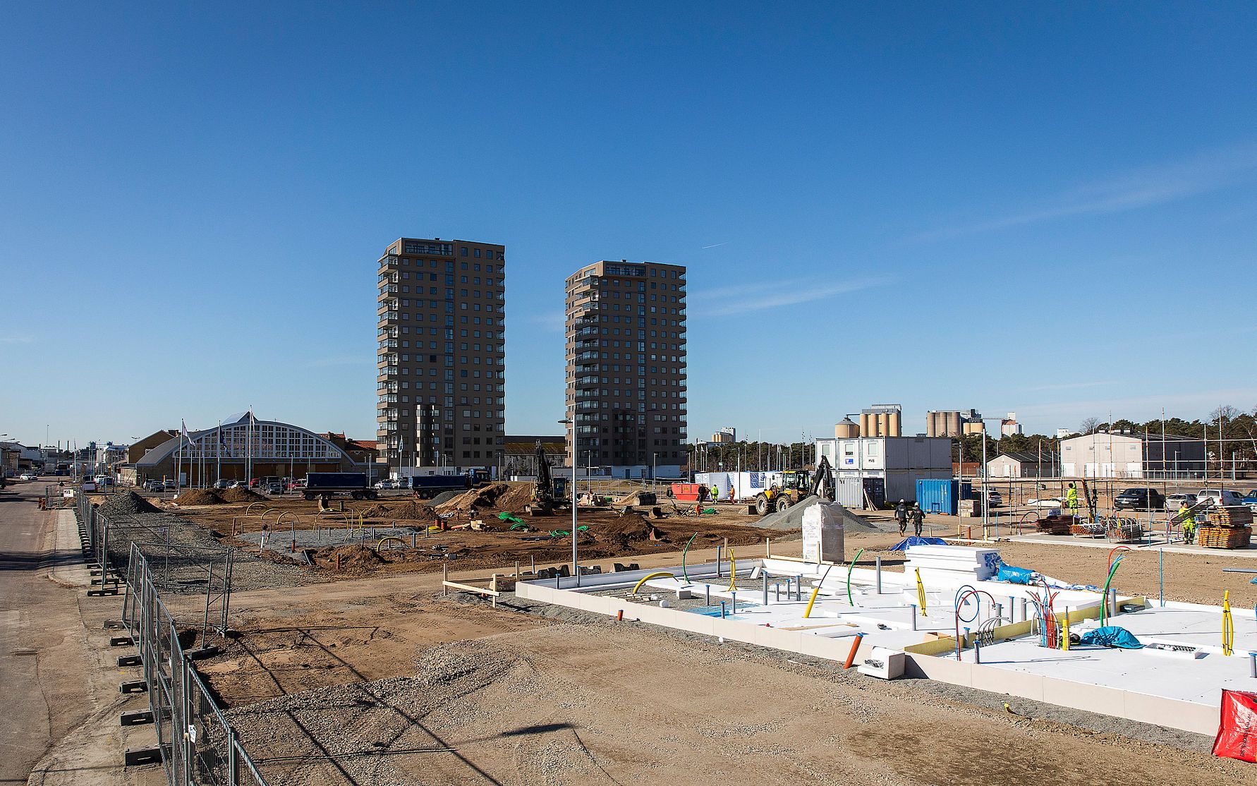 Byggarbetsplats med flerbostadshus i bakgrunden, byggmaterial och utrustning på marken under en klar blå himmel