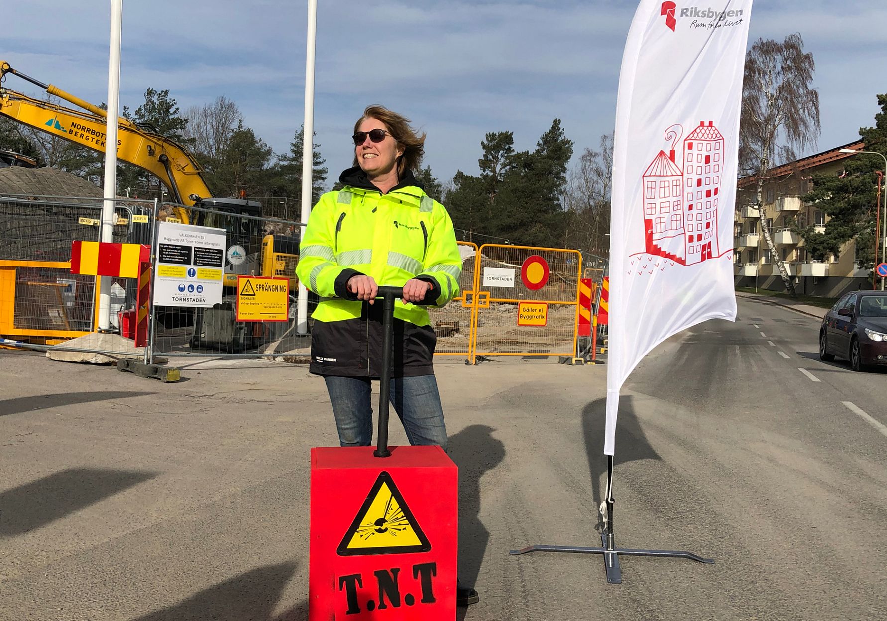Person i en gul reflexjacka står på en väg med en röd T.N.T-markör framför sig. I bakgrunden syns byggarbetsplats, avspärrningsskyltar och en flagga med en logotyp