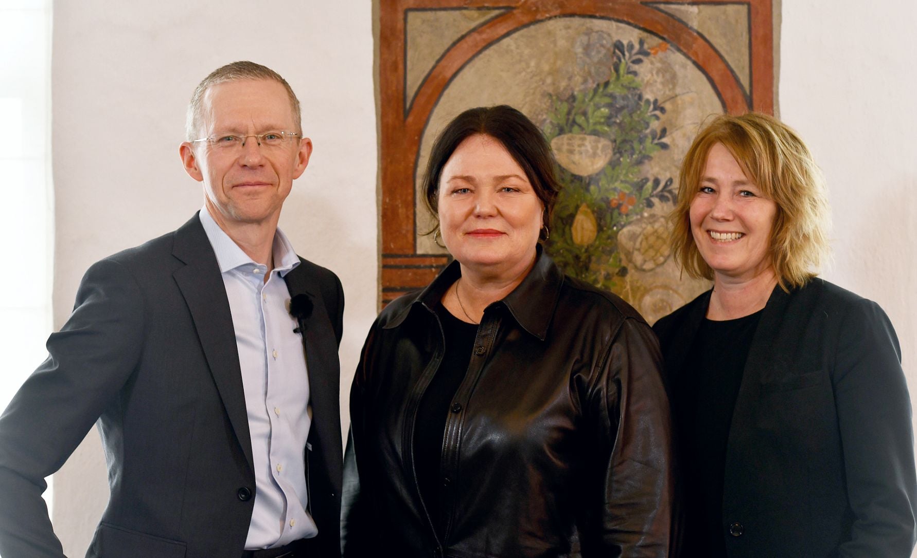 Tre personer står framför en målning med växtmotiv, iförda formella kläder