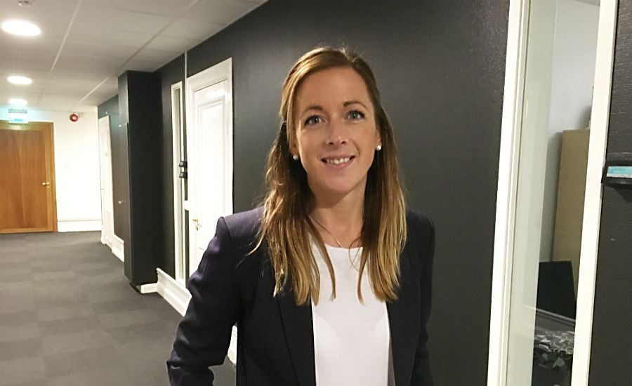 Kvinna i kavaj och vit topp står i en kontorskorridor med vita dörrar och mörka väggar