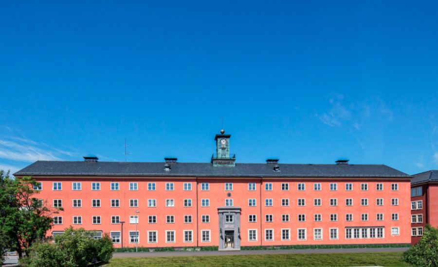 Röd byggnad med flera fönster och en klocka på taket, omgiven av gräsmatta och blå himmel