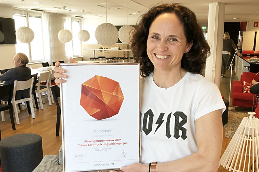 Kvinna i vit t-shirt håller i en utmärkelse med text och en grafisk design, inuti en modern inredd lokal med bord och stolar