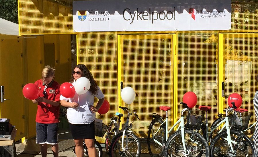 En cykelpool med gula metallväggar i bakgrunden, flera uppställda cyklar och två personer som håller i röda och vita ballonger