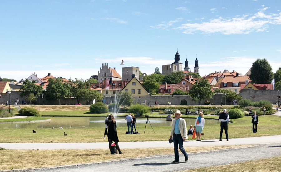 Stadsvy med grönområde, fontän och människor som promenerar, med äldre byggnader och kyrktorn i bakgrunden under en klarblå himmel