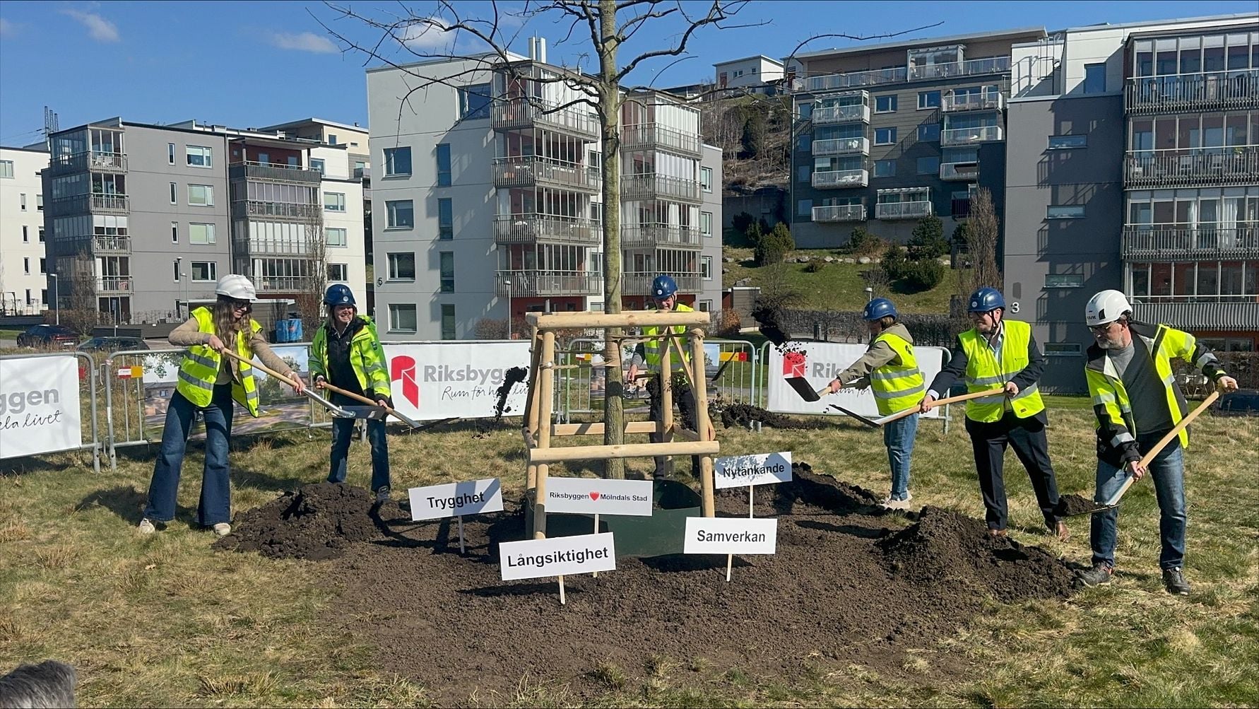 Grupp av personer i reflexvästar och hjälmar som tar ett första spadtag på en byggplats med flerbostadshus i bakgrunden