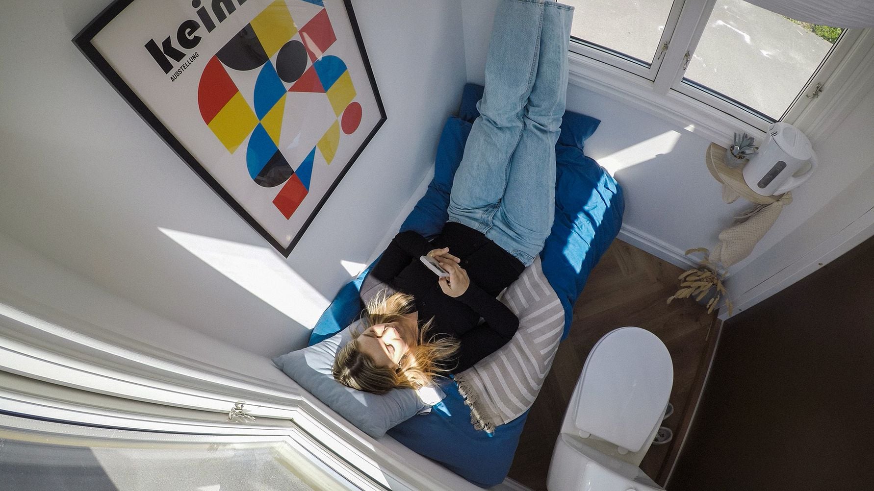 Person liggande på bäddsoffa i ett ljust rum med fönster, mobiltelefon och väggkonst