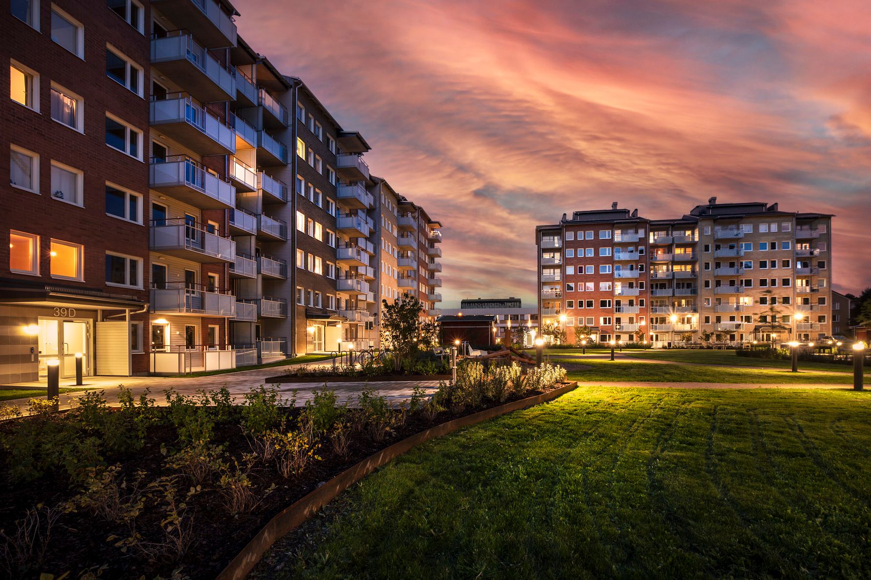 Flerbostadshus med moderna fasader och balkonger vid solnedgång, omgivna av grönområden och välskötta planteringar