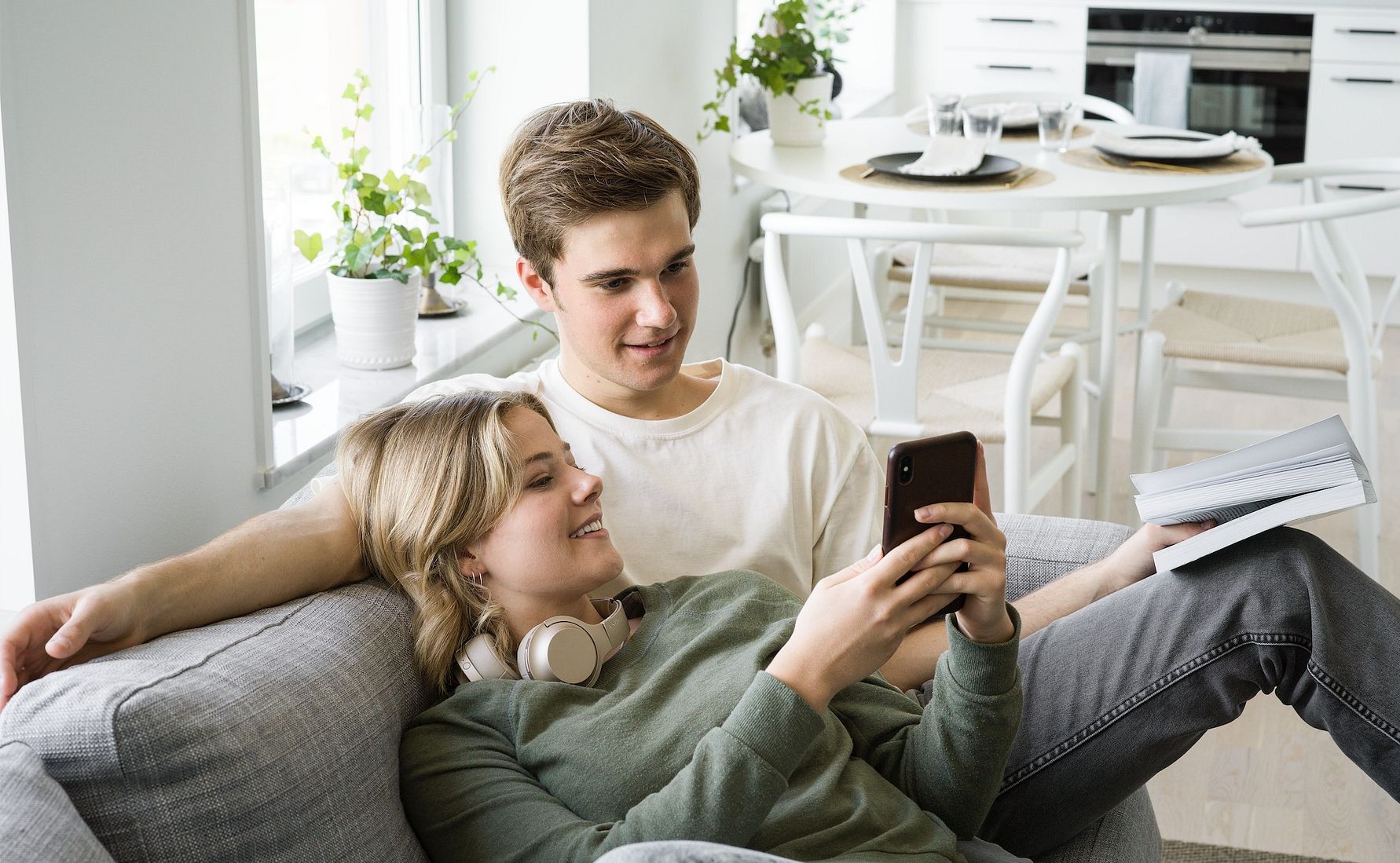 Ett ungt par sitter i en modern och ljus vardagsrumsmiljö, där den ena personen håller en smartphone och den andra håller en bok