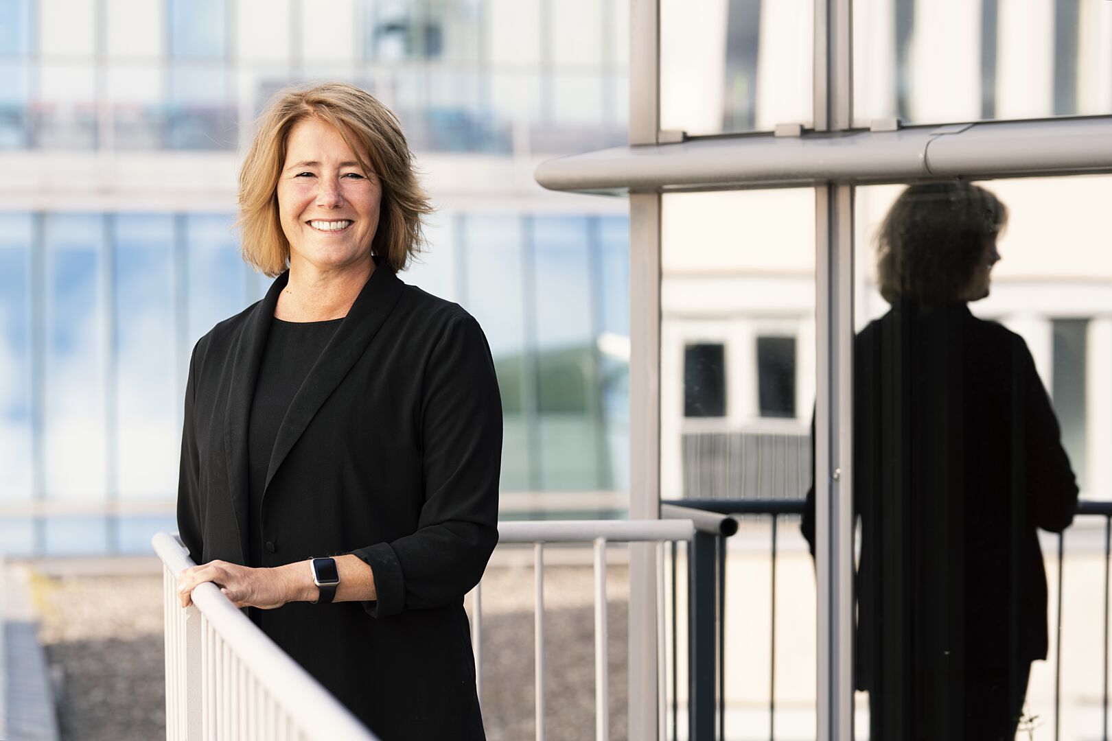 Porträtt av Riksbyggens VD Johanna Frelin i svart klädsel som står vid ett räcke, med sin spegelbild i en glasvägg