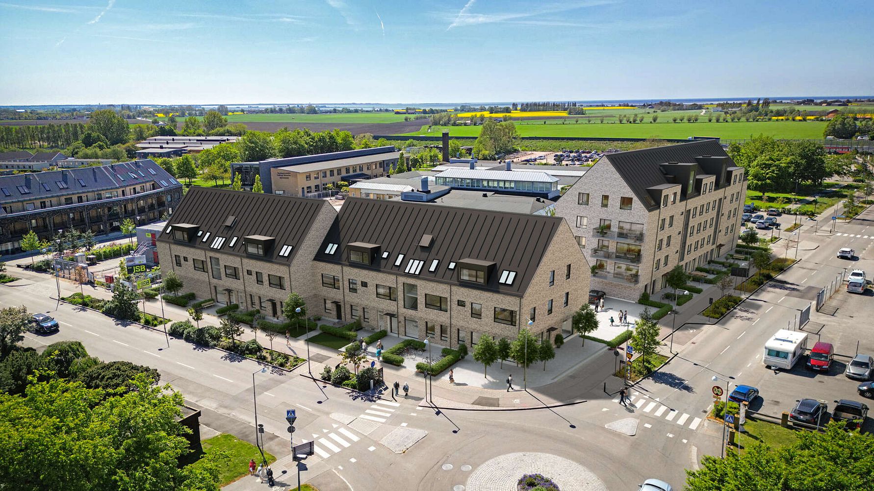 Vy över ett modernt bostadsområde med flerfamiljshus och radhus, omgiven av grönska och öppna fält under klarblå himmel