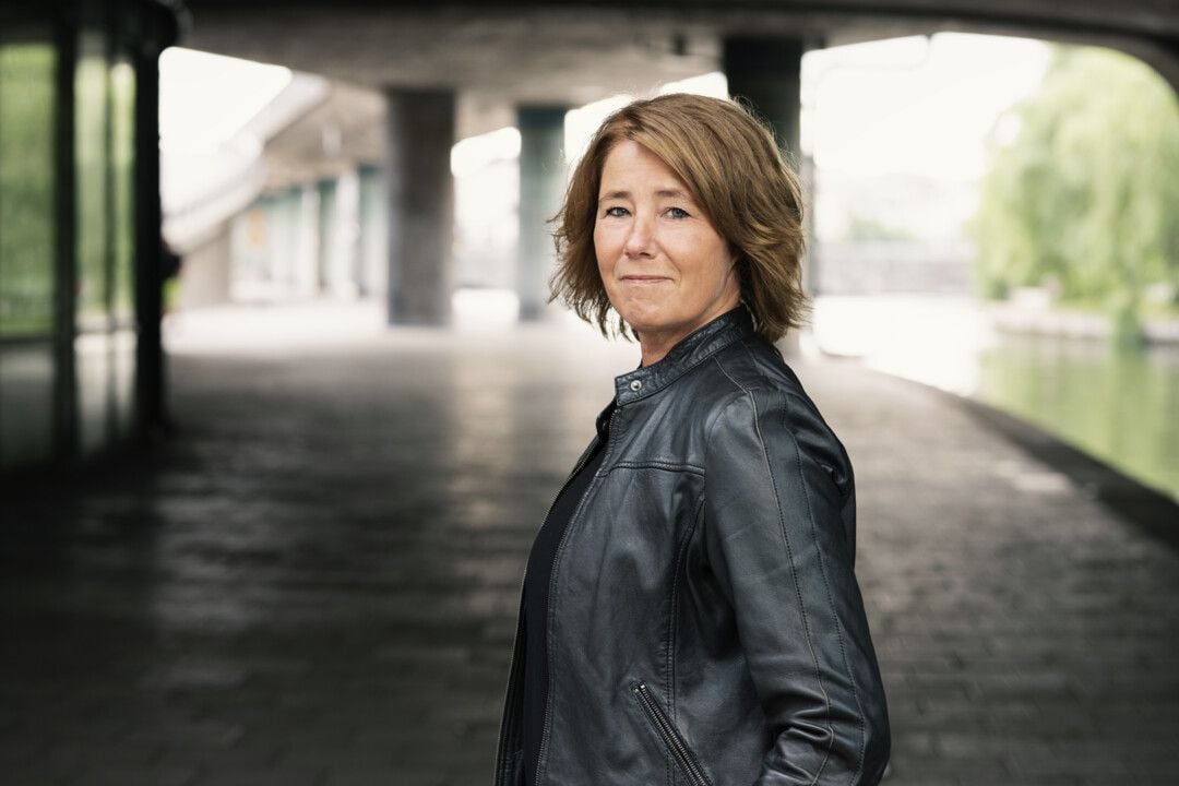 Kvinna i svart läderjacka står under en bro i en urban miljö med gångväg och vatten i bakgrunden