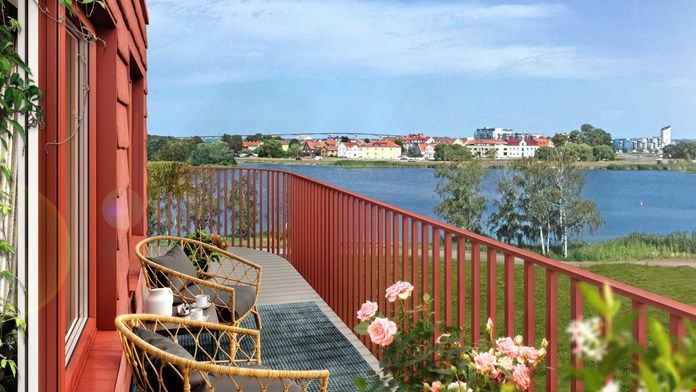 Utomhus balkong med röda räcken, två stolar och bord, blommor i förgrunden, med utsikt över en sjö och bostadshus i bakgrunden