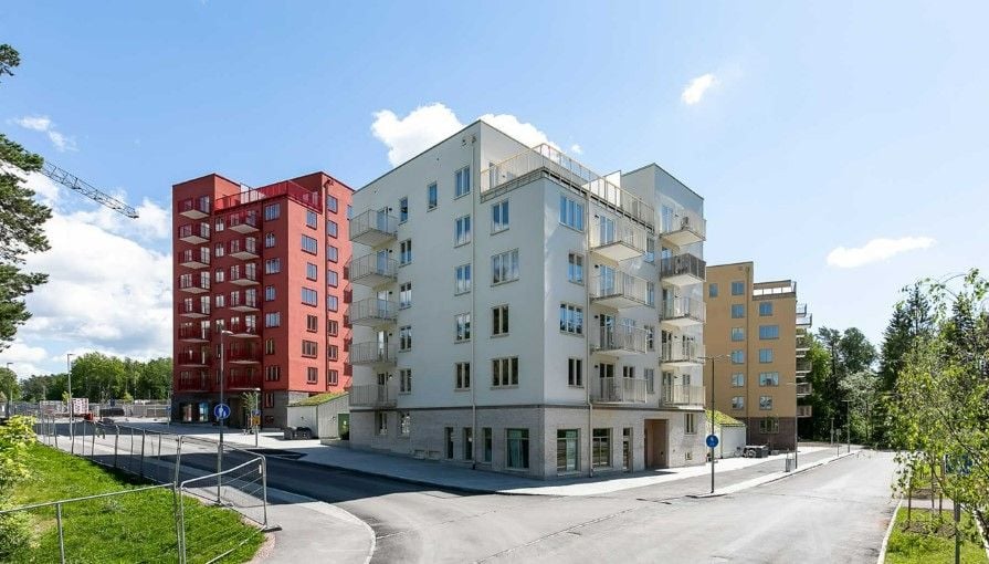 Flera moderna flerfamiljshus i olika färger på en solig dag, omgivna av grönska och en asfalterad väg