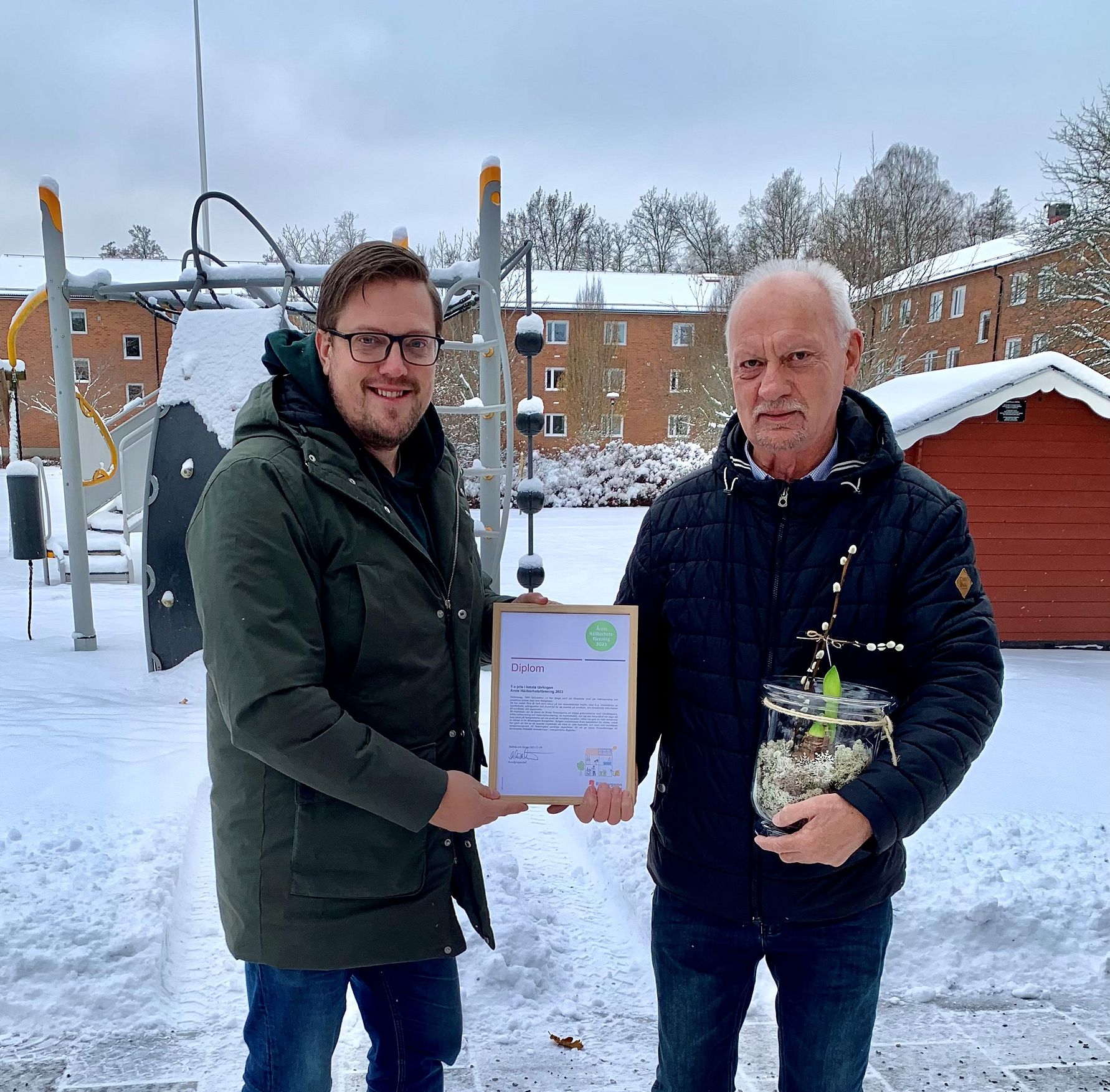 Två personer står utomhus i en snöig miljö, den ena håller ett diplom och den andra håller en blomsterarrangemang i en glaskruka. Bakgrunden visar en lekplats, flerbostadshus och snötäckta träd