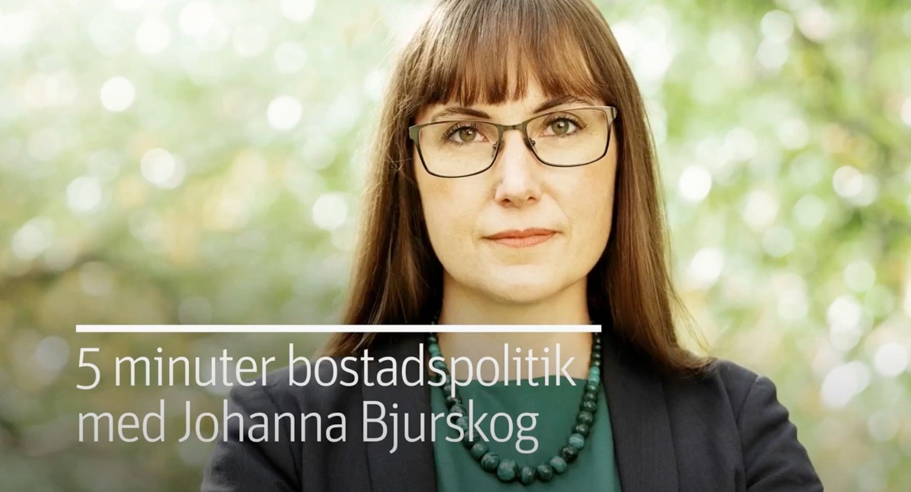 En person i formell klädsel med ett halsband, stående utomhus framför en grönskande bakgrund. Texten på bilden lyder "5 minuter bostadspolitik med Johanna Bjurskog