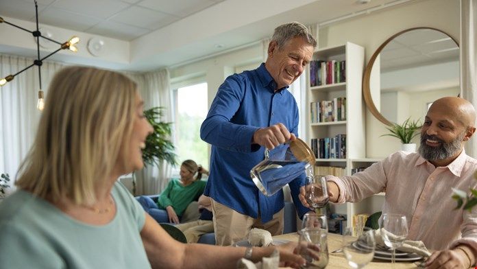 En grupp människor sitter vid ett dukat matbord i ett ljust rum, där en person häller vatten i ett glas