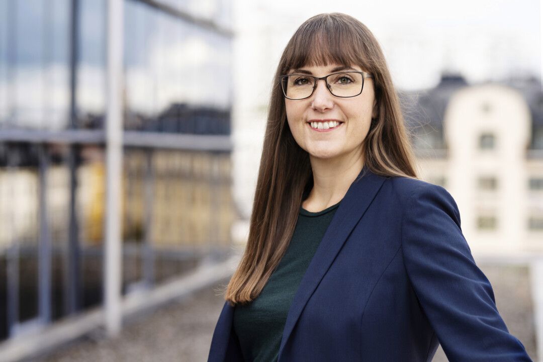Kvinna i mörkblå kavaj och svart topp står framför en modern byggnad med glasfasad och stadsmiljö i bakgrunden