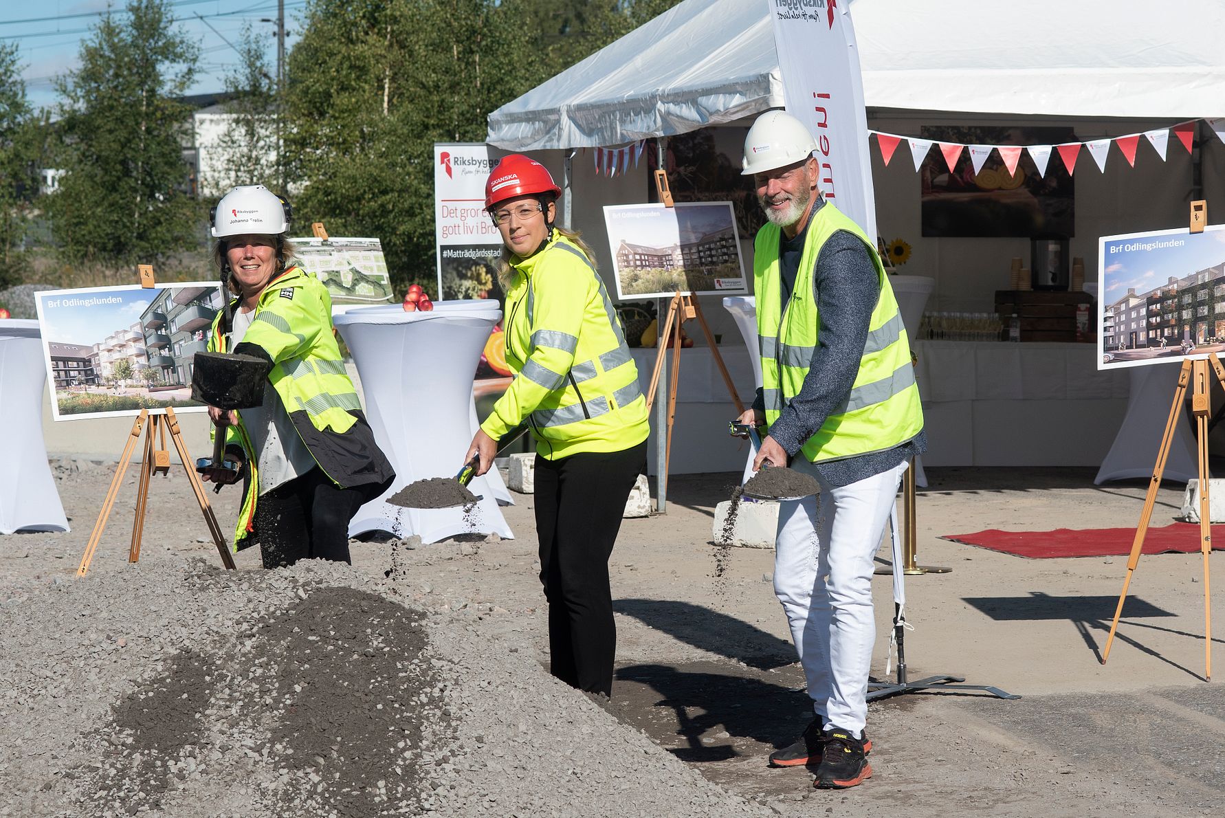 Tre personer i reflexvästar och hjälmar skyfflar sand vid en ceremoni på en utomhusplats, med tält och informationsskyltar i bakgrunden
