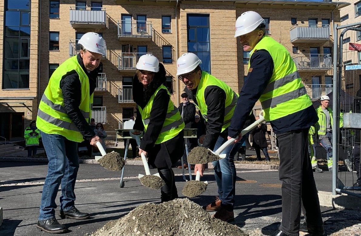 Fyra personer i reflexvästar och byggarbetsplats-hjälmar skyfflar grus med spadar vid en byggarbetsplats, med en flerbostadsbyggnad i bakgrunden