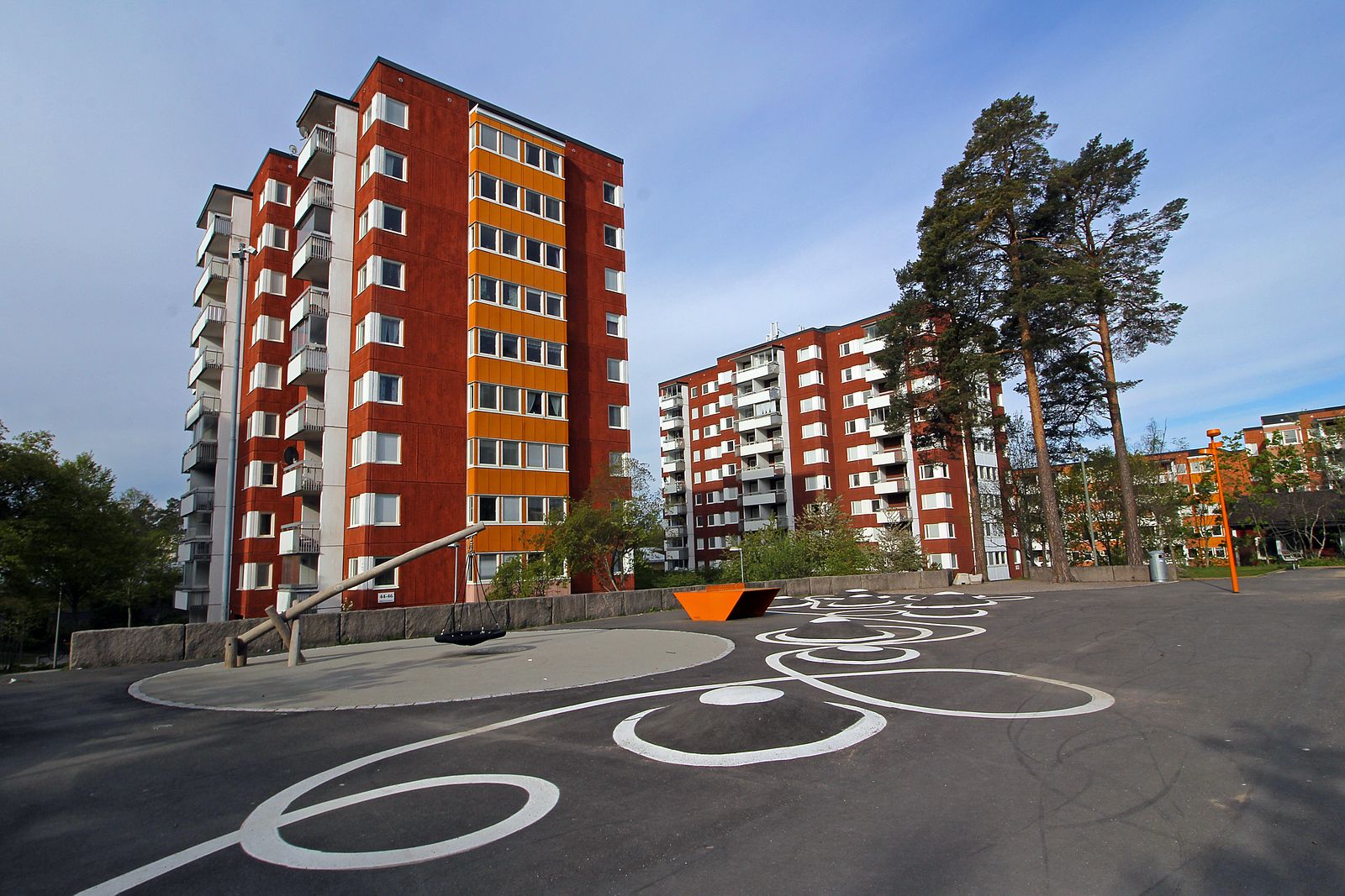 Flerbostadshus med röda fasader och vita balkonger, omgivna av träd och en lekplats med asfaltmönster