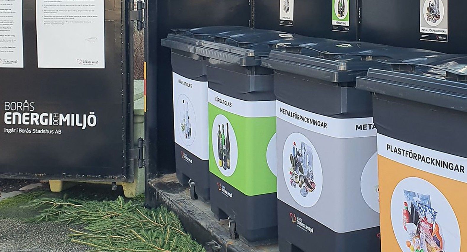 Återvinningsstation med soptunnor för plastförpackningar, metallförpackningar och blandat avfall, omgiven av grönska. Texten "Borås Energi och Miljö" synlig på en skylt