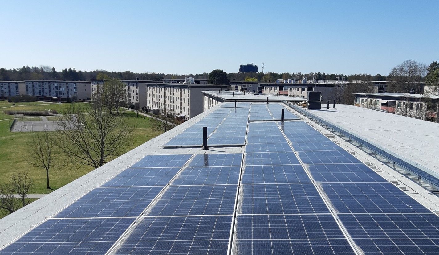 Solpaneler på taket av en byggnad med bostadshus och grönområden i bakgrunden under klarblå himmel