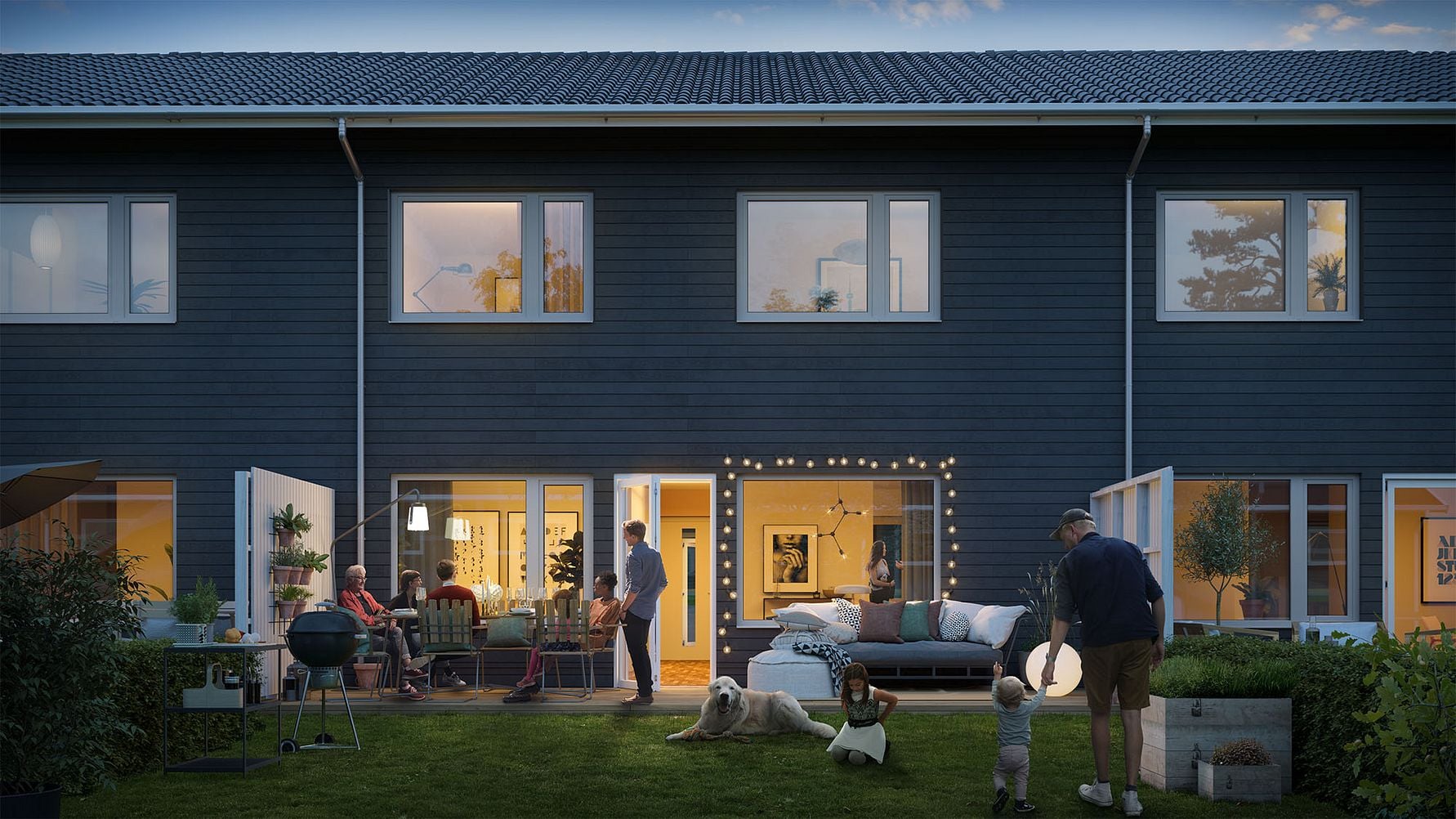 Tvåvåningshus med mörk fasad på kvällen, med människor som umgås i trädgården och interiören upplyst