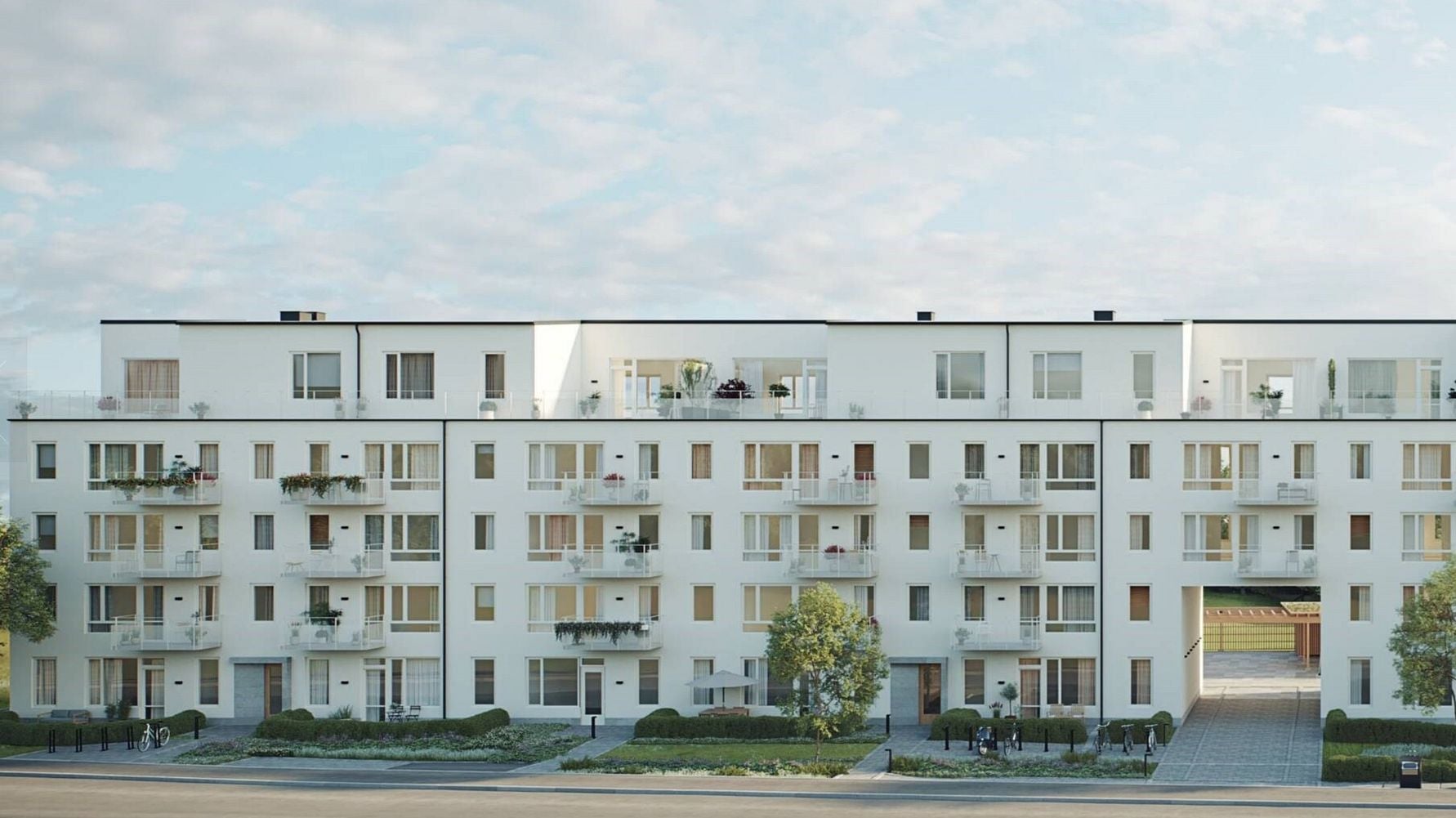Vit flerfamiljsbyggnad med balkonger och fönster, omgiven av grönska och trottoar under en ljus himmel