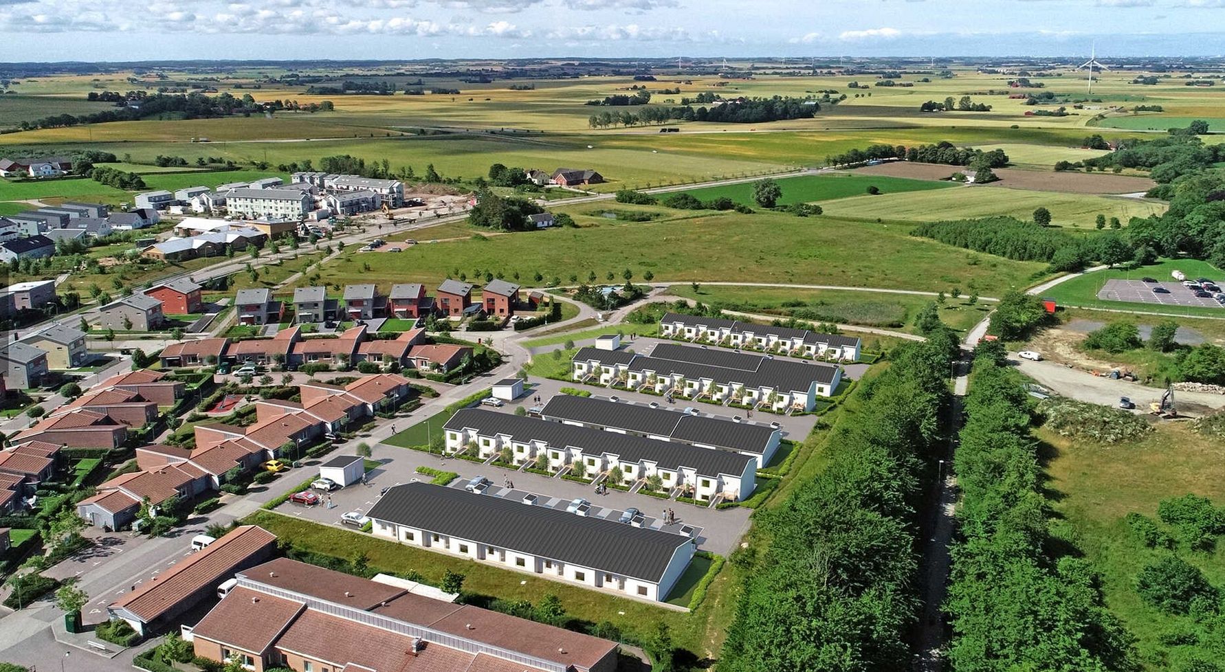 Flygbild av en förort med radhus, gröna fält och lantlig omgivning under en klar himmel