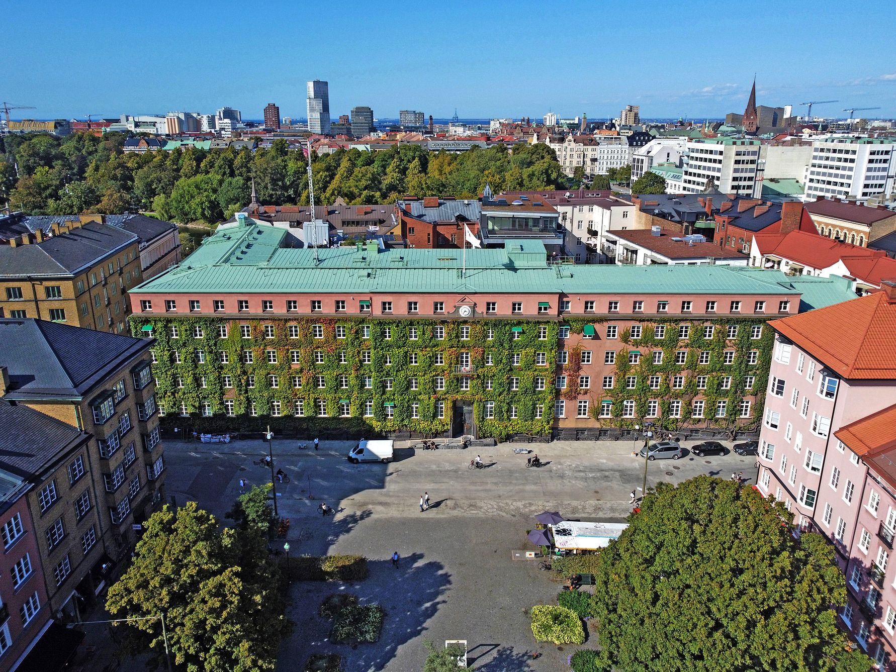 Flygfoto av en stor tegelbyggnad med grön växtvägg, omgiven av träd och stadsmiljö, med en klar blå himmel i bakgrunden