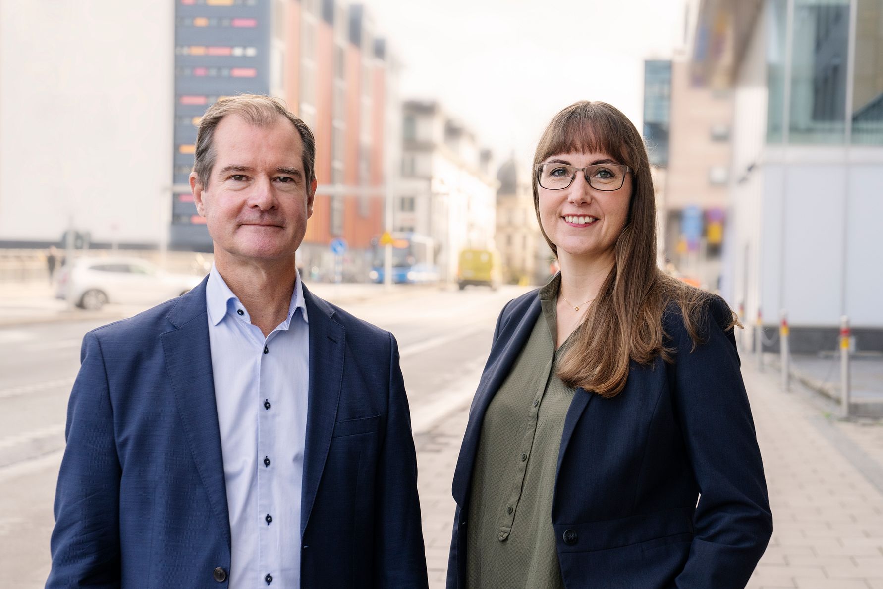 Två personer i affärskläder står utomhus på en stadsgata med moderna byggnader i bakgrunden