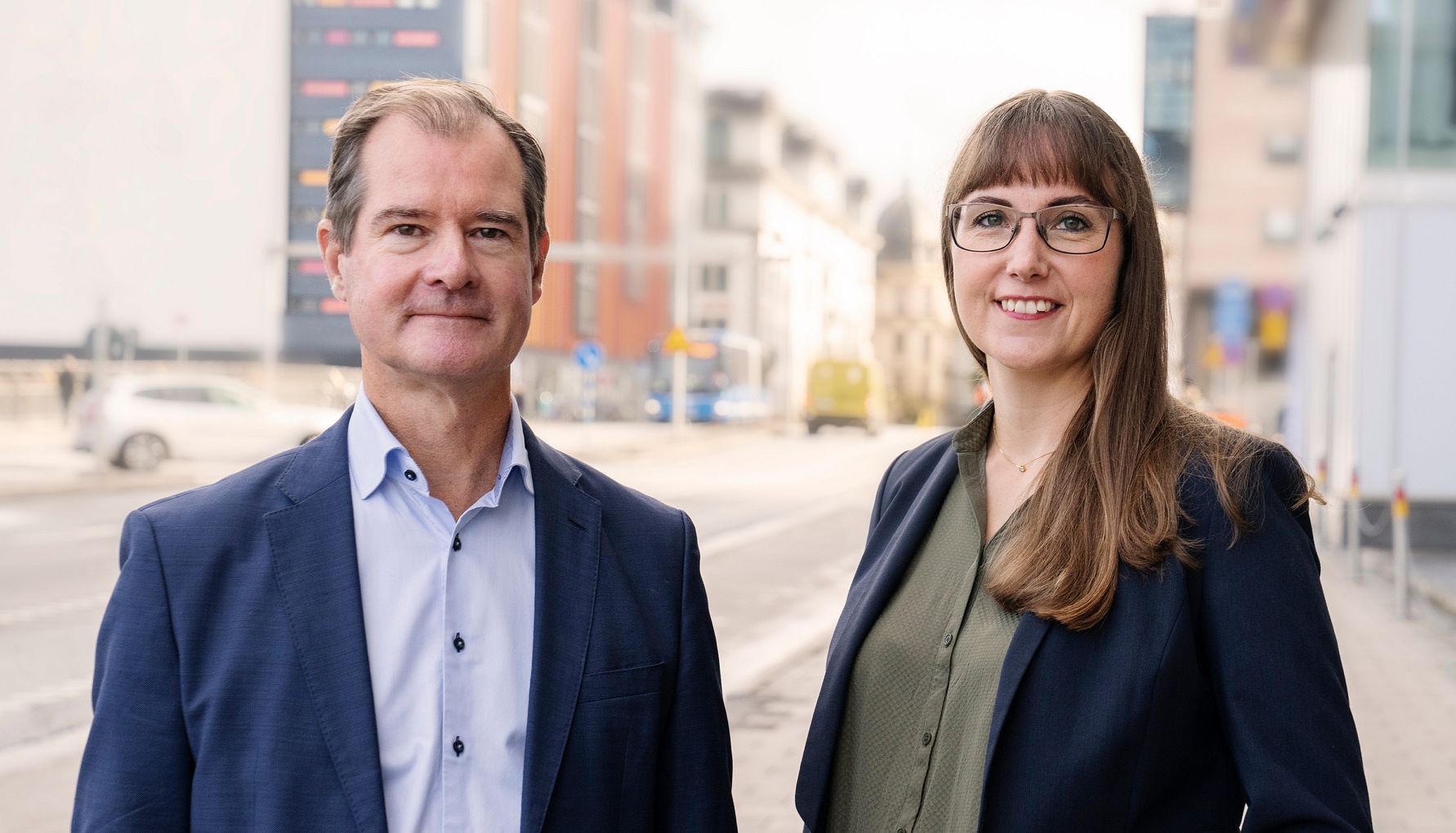 Två personer i formella kläder står utomhus på en gata med moderna byggnader i bakgrunden