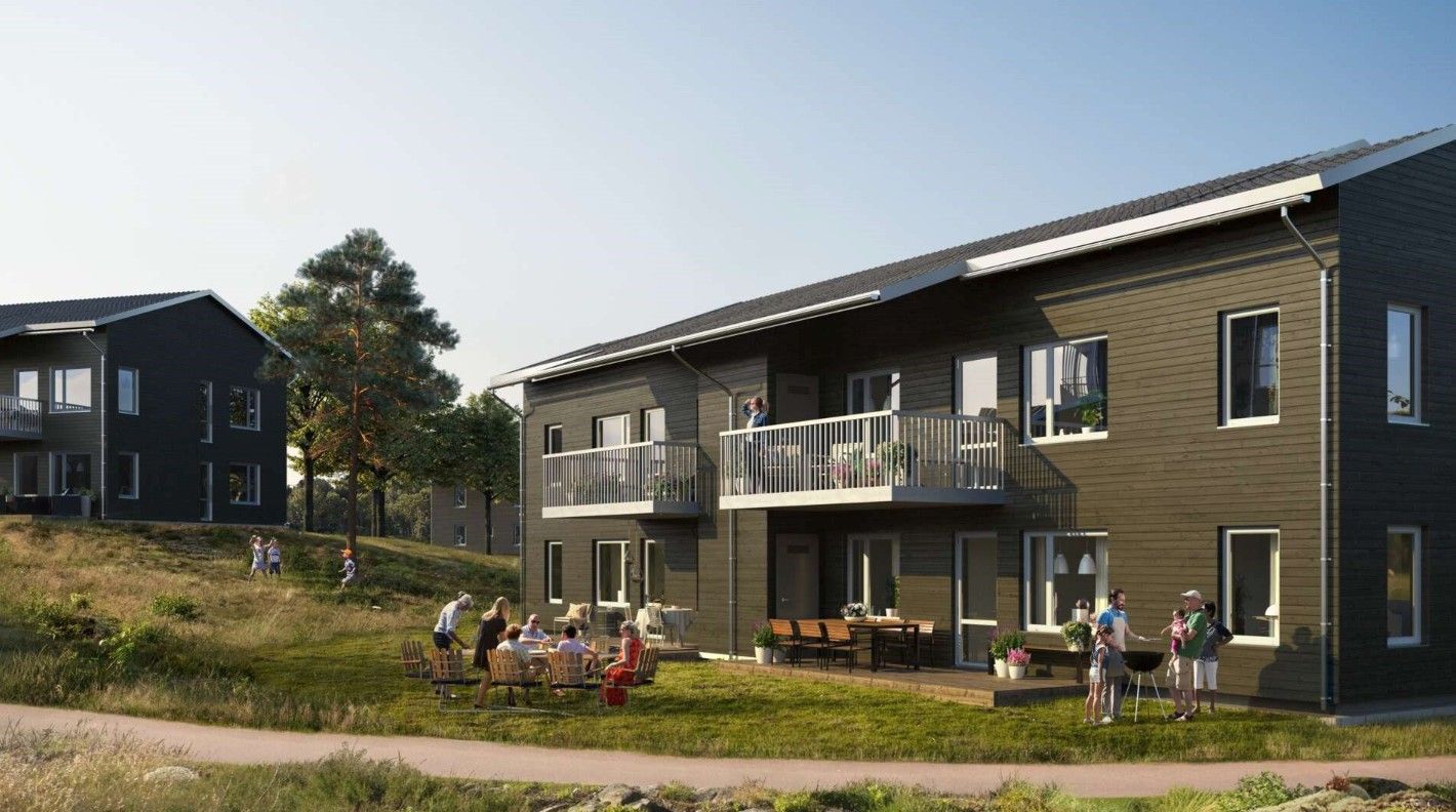 Två moderna, mörka trähus med solpaneler, omgivna av grönska. Människor umgås utomhus vid en bordgrupp och på balkonger under en solig dag