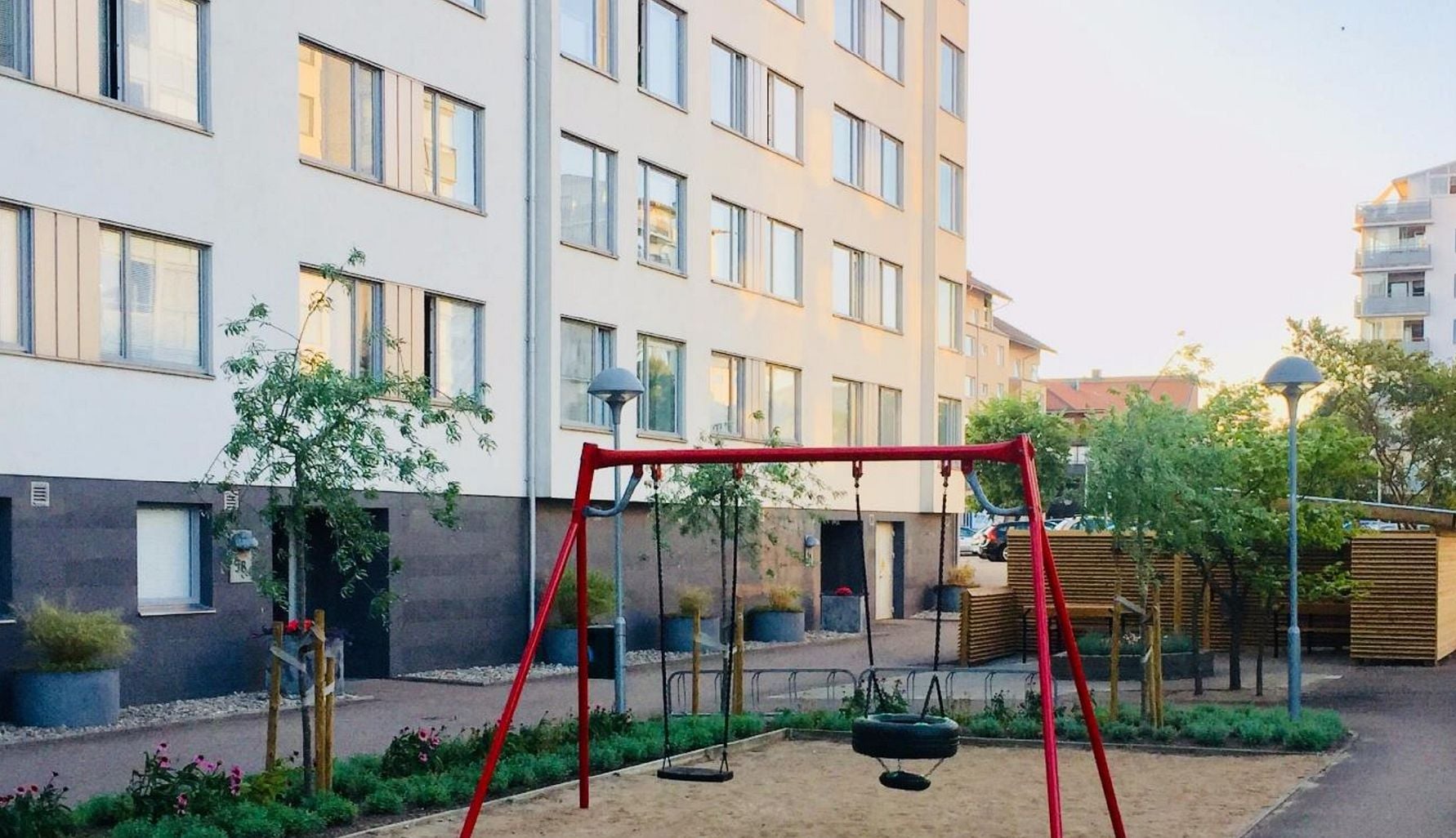 Lekplats med gungställning framför ett flerbostadshus i ett bostadsområde, omgiven av grönska och planteringar