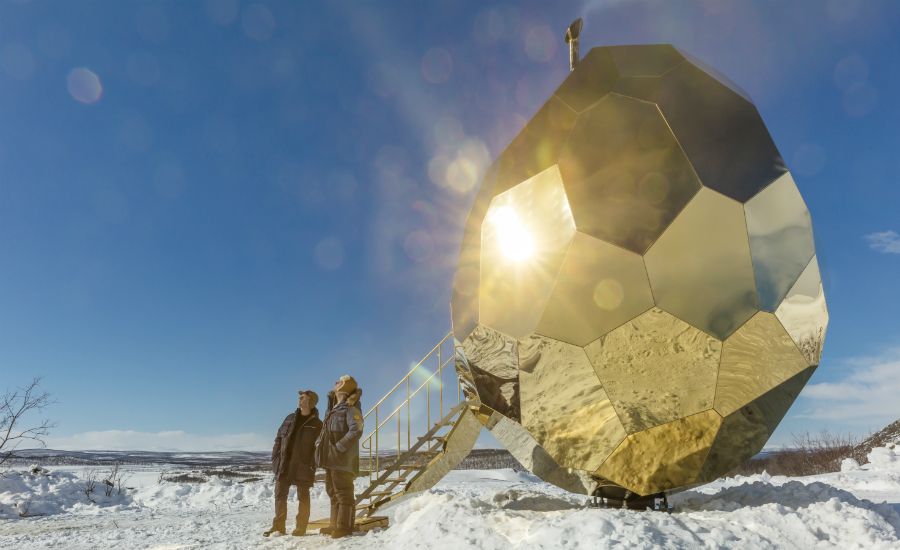 Två personer står framför en stor, guldfärgad geometrisk byggnad i ett snötäckt landskap under klar blå himmel