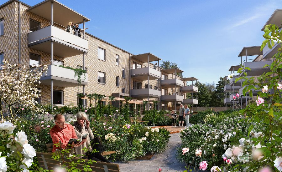 Modernt bostadsområde med flerfamiljshus i tegel, omgiven av trädgårdar med blommor och gröna buskar. Människor sitter på bänkar och promenerar i det soliga området