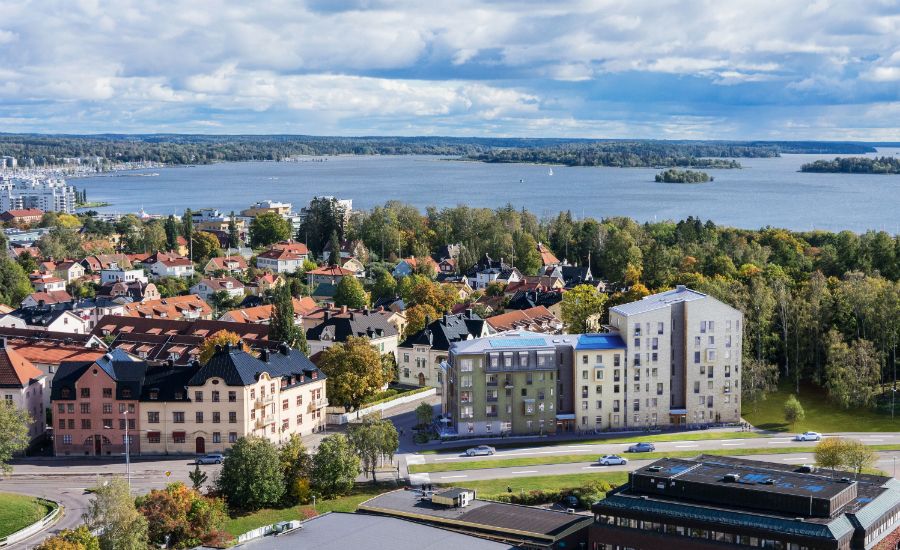 Flygfoto över ett bostadsområde med flerfamiljshus, gröna träd, och en sjö i bakgrunden under en molnig himmel
