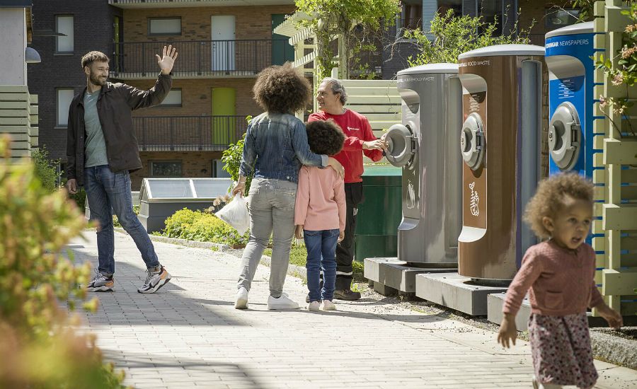 En grupp människor på en promenad nära återvinningsstationer i ett bostadsområde med grönska och flerbostadshus i bakgrunden