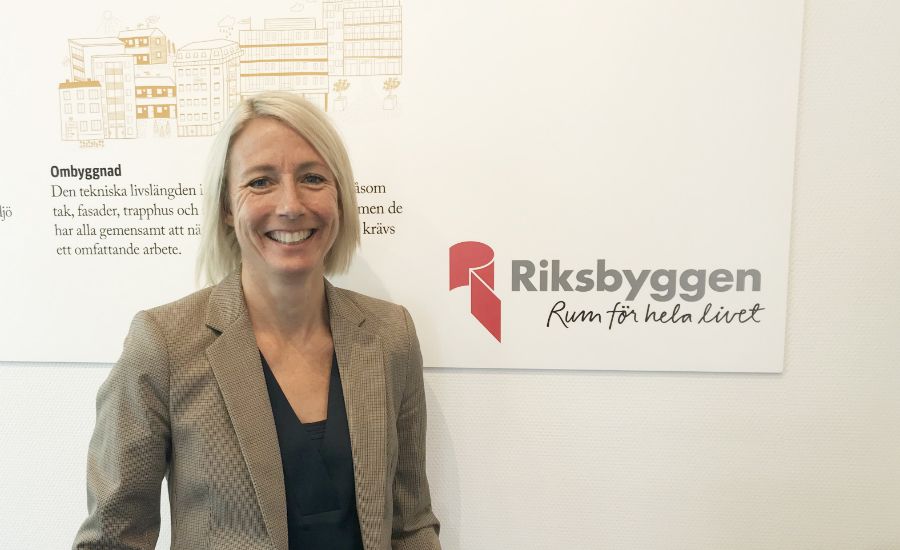 Kvinna i kavaj står framför en vit vägg med en affisch som visar text och illustrationer samt logotypen för Riksbyggen och texten "Rum för hela livet