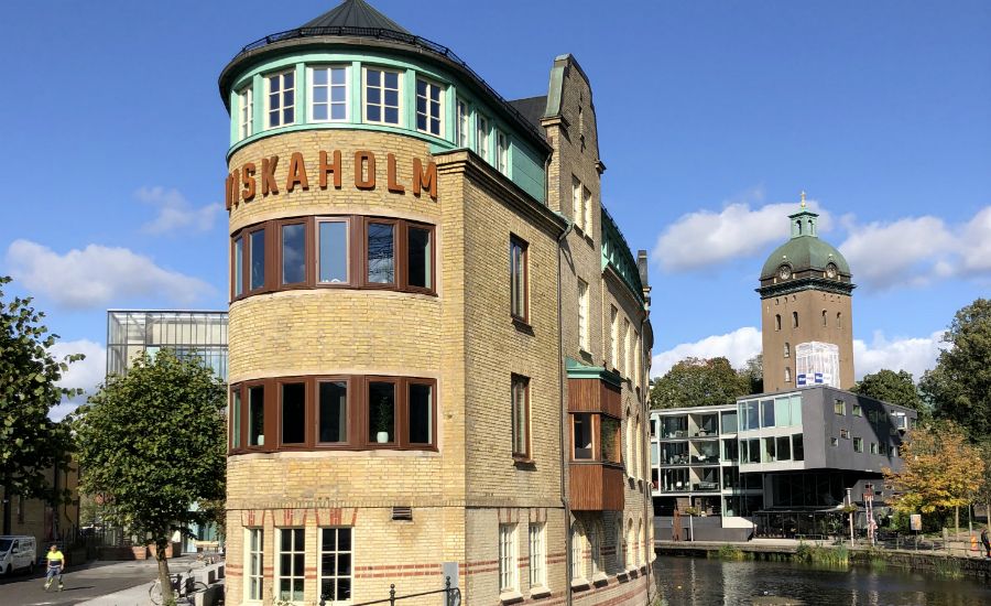 Gul tegelbyggnad med texten "ARISEKAHOLM" nära en kanal, omgiven av grönska och modern arkitektur i bakgrunden under en klarblå himmel